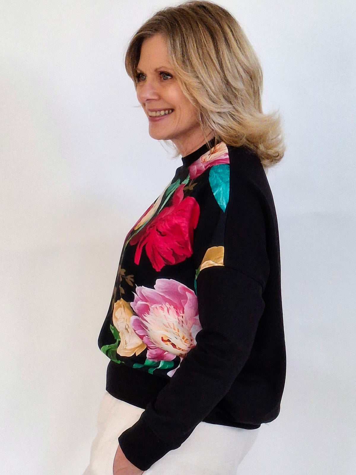 A woman with blonde hair is shown in profile wearing a black knit sweater with a vibrant multicolor floral pattern on the front and white pants, set against a plain white studio background.