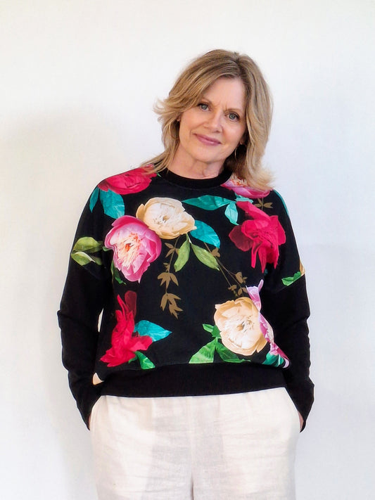 A woman wearing a black crewneck sweatshirt with a vibrant floral print of pink, red, and cream roses with green leaves, paired with light beige trousers, set against a plain white background.